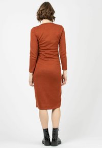 Robe ajustée à manches longues de couleur rouille, avec une longueur au genou. Fabriquée à partir d'un matériau doux. Portée avec des bottines noires à la cheville.