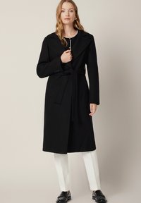 Czarny płaszcz z mieszanki wełny z paskiem w talii, dużym kołnierzem i długości do kolan, w połączeniu z białymi spodniami i czarnymi butami.