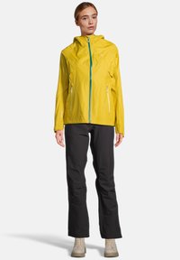 Geel lichtgewicht jack met een capuchon, ritssluiting aan de voorkant en teal accenten. Voorzien van ritszakken en een ademende, soepele textuur. Zwarte broek maakt de look af.