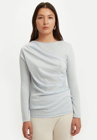 Hellblaues Langarm-Oberteil aus weichem, strukturiertem Stoff mit drapiertem Design und gerafften Details an der Seite. Rundhalsausschnitt.