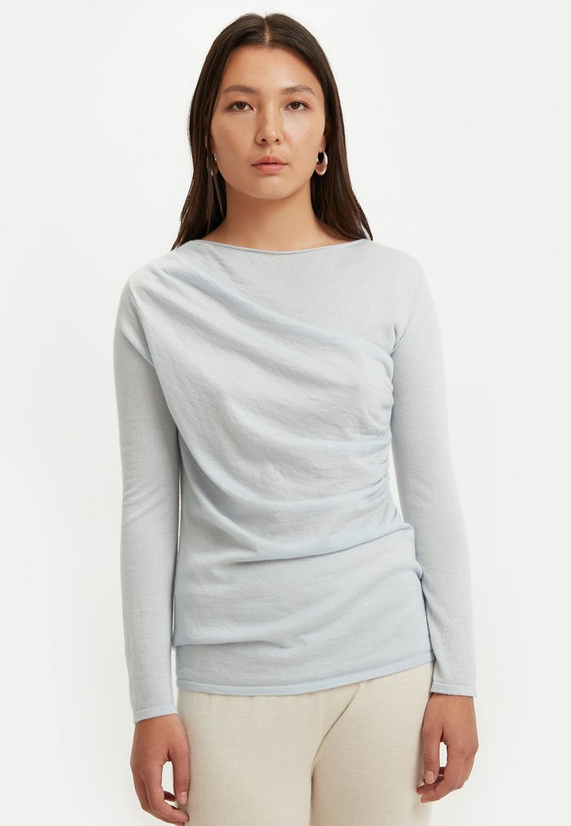 Hellblaues Langarm-Oberteil aus weichem, strukturiertem Stoff mit drapiertem Design und gerafften Details an der Seite. Rundhalsausschnitt.
