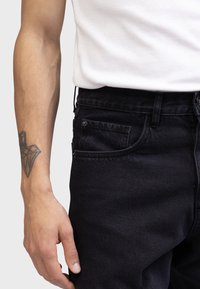 Jeans negri din denim cu o textură netedă, având un buzunar frontal, bucle pentru curea și un top tip tank alb, ajustat. Se vede un braț tatuat.