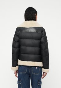 Eine Frau von hinten zu sehen, die eine schwarze Daunenjacke mit beigem Schafslederkragen und -manschetten trägt, kombiniert mit blauen Jeans-Cargo-Hosen.