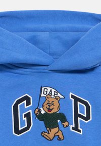 Felpa con cappuccio blu con grandi lettere nere e bianche "GAP" e un orsetto dei cartoni che tiene una bandiera bianca con il testo "GAP".