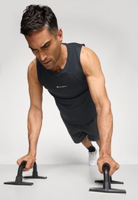 Schwarzes Sporttop und Shorts, in der Liegestützposition mit schwarzen Liegestützgriffen auf einer hellen Oberfläche. Neutraler Hintergrund.