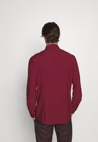 Camisa de manga larga color burdeos con cuello, puños abotonados y corte entallado. La tela parece suave con un ligero brillo.