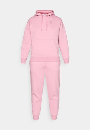 Conjunto de sudadera con capucha y pantalones deportivos en suave tejido, que incluye un bolsillo tipo canguro, capucha con cordón, y un pequeño parche con logo en el pecho y el muslo.