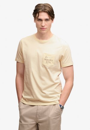 Junger Mann mit hellbraunen Haaren, der ein beiges Superdry-T-Shirt und Khakihosen trägt, mit den Händen in den Taschen vor weißem Hintergrund.