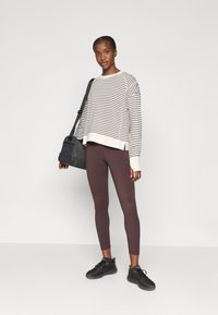 Gestreifter langärmliger Sweatshirt in Creme und Dunkelbraun, kombiniert mit bordeauxfarbenen Leggings und schwarzen Turnschuhen. Enthält eine schwarze Umhängetasche.
