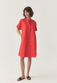 Red cotton dress with short sleeves, front button closure, and side pockets. Straight cut, knee-length with a smooth texture. Black slide sandals.