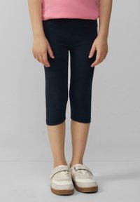 Leggings bleu marine en tissu extensible, coupés au niveau du mollet. Portés avec des baskets beiges dotées d'une sangle Velcro et d'une semelle brun clair.