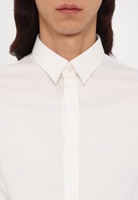 Gros plan d'une personne portant une chemise blanche boutonnée avec col, avec de longs cheveux foncés visibles sur les côtés.