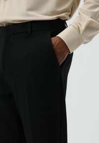 Zwarte broek met een getailleerde pasvorm, voorzien van een zijzak en een plooi langs het been. Draagt een lichtbeige, gestructureerd shirt met lange mouwen.