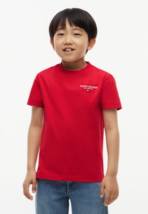 Νεαρός αγόρι με κόκκινο T-shirt Tommy Hilfiger και μπλε τζιν, στέκεται μπροστά σε λευκό φόντο, χαμογελώντας ελαφρώς.