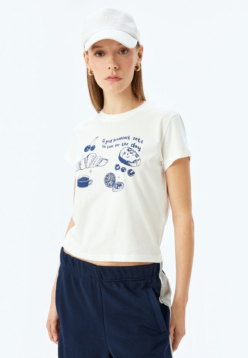 Witte katoen t-shirt met marineblauw ontbijtthema grafiek, featuring croissant, kopje en fruit. Gekorte ontwerp met opgerolde mouwen.
