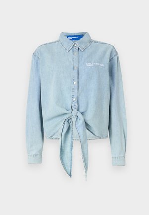 Camicia in denim azzurro chiaro con colletto a punta e maniche lunghe. Presenta un dettaglio di nodo legato in vita e un logo ricamato sul petto.