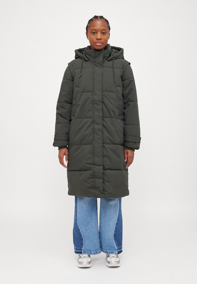 Olivengrønn dunjakke med glidelås foran, justerbar hette og sidelommer. Brukt over lyseblå bredbeinte jeans og sneakers.