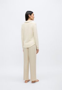 Bézs színű pizsama szett hosszú ujjakkal és bő szárú nadrággal. Puha, texturált anyagból készült. Galléros nyakkivágással és laza szabással rendelkezik.