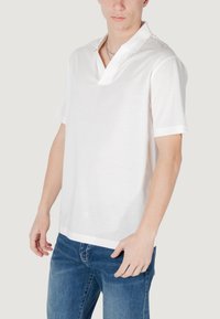 Polo bianca con colletto corto, realizzata in cotone leggero. Presenta maniche corte e una vestibilità comoda, indossata con jeans blu.