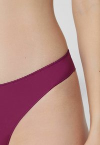 Bikini dolny w głębokim śliwkowym kolorze, o gładkiej fakturze, niskim kroju oraz przeszyciach wzdłuż brzegów, zapewniający precyzyjne dopasowanie.