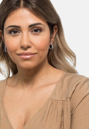Blouse beige à col en V avec une texture douce. Boucles d'oreilles carrées en argent avec un motif texturé. Le mannequin a des cheveux mi-longs et ondulés et un maquillage neutre.