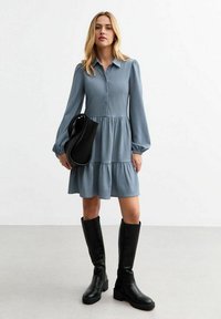 Blue button-up dress with a textured fabric, balloon sleeves, and a tiered skirt. Worn with black knee-high boots and a black bag.