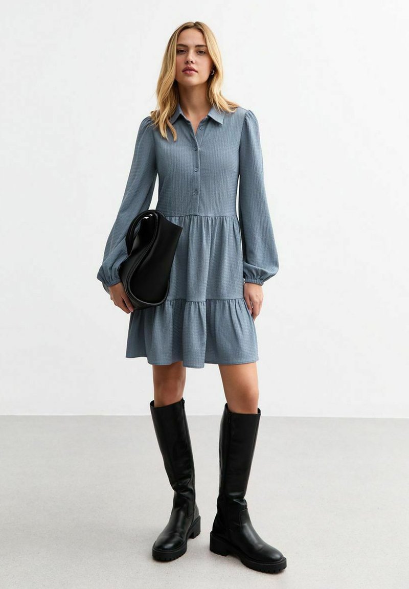 Blue button-up dress with a textured fabric, balloon sleeves, and a tiered skirt. Worn with black knee-high boots and a black bag.