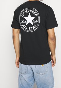 Osoba viděná zezadu, nosící černé tričko s velkým bílým logem Converse All Star, které obsahuje hvězdu a text. Světle modré džínové kalhoty.