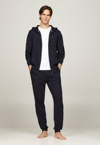 Marineblaue Zip-Hoodie mit Kordelzug, Fronttaschen und einem weißen T-Shirt darunter, kombiniert mit passenden marineblauen Sweatpants, minimalistisches Design, weiche Textur.