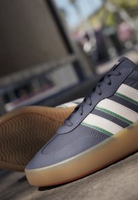 Zapatilla Adidas azul y blanca con suela de goma, con detalles verdes y cierre de cordones en la parte delantera, colocada al aire libre sobre el pavimento.