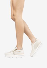 Beige sneakers with leather and mesh panels, featuring a zipper, metallic accents, and a white sole. Eased fit for comfort.