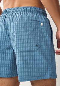 Sinisellä ruutukuvioidusta teksturoidusta kankaasta valmistetut uimashortsit. Niissä on joustava vyötärönauha, sivutaskut ja takatasku napillisen suljettavan läpän kanssa.