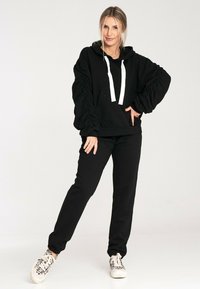 Czarna bluza z kapturem z białymi sznurkami, z marszczonymi rękawami i przednią kieszenią, zestawiona z dopasowanymi czarnymi spodniami i wzorzystymi białymi trampkami.