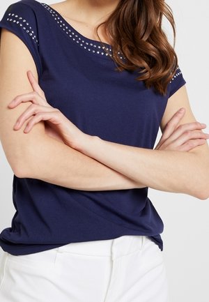 Vrouw draagt een marineblauw shirt met korte mouwen en een met studs versierde halslijn, armen gekruist, gecombineerd met een witte broek tegen een effen achtergrond.