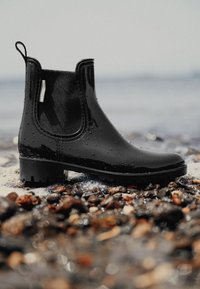 Zwarte waterdichte Chelsea boot van glanzend materiaal, met elastische panelen en een lage gestapelde hak, met waterdruppels op het oppervlak.