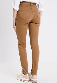 Pantalon ajusté marron avec une taille haute, des poches arrière et une finition lisse, associé à des baskets beiges avec des accents métalliques.