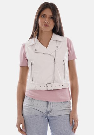 Giovane donna che indossa un gilet bianco senza maniche con cerniera e cintura sopra una maglietta rosa a maniche corte e jeans azzurri.