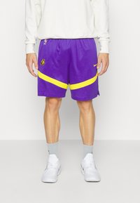 Lila basketshorts med gula detaljer och ränder, med en broderad logotyp och meshmaterial. Bärs med grå strumpor och vita sneakers.