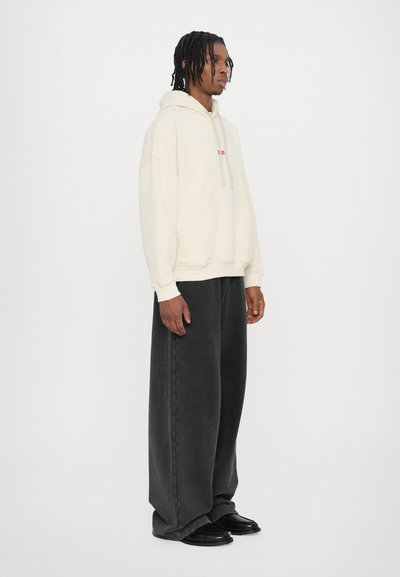 Cream oversized hoodie with front pocket and red text, paired with wide-leg dark gray pants. Model wears black shoes.