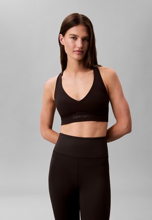 Kobieta w czarnym sportowym biustonoszu i legginsach z wysokim stanem stoi ze złożonymi rękami za plecami na tle gładkiej szarej ściany.