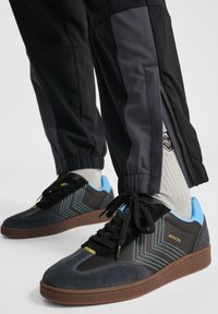 Svarta sneakers med blå detaljer och zigzag-mönster; i mocka och syntetmaterial; gummisula; kombineras med svarta byxor med dragkedja vid fållarna.