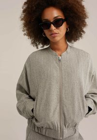 Jachetă bomber gri deschis cu model în herringbone, închidere cu fermoar, manșete și tiv elastic. Modelul poartă ochelari de soare negri și cercei cerc.