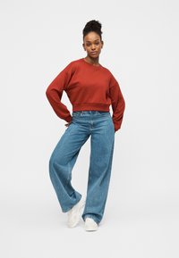 Κοντοκομμένο κόκκινο φούτερ με μακριά μανίκια, συνδυασμένο με ανοιχτόχρωμα τζιν φαρδιάς γραμμής και λευκά αθλητικά παπούτσια· απλός, καθημερινός σχεδιασμός.