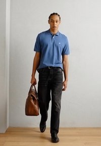 Cămașă polo albastră, blugi negri, geantă din piele maro. Modelul are părul împletit și poartă pantofi negri. Design simplu și paletă de culori neutră.