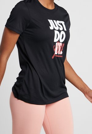 Personne portant un t-shirt noir avec le texte "JUST DO IT." et des leggings rose clair, montrée des épaules aux hanches sur un fond uni.