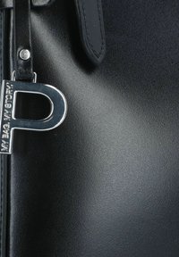 Schwarze Handtasche mit strukturierter Oberfläche, die einen silbernen Metallscharm in Form des Buchstabens "P" und einen glatten Reißverschlusszug mit Lederakzenten aufweist.