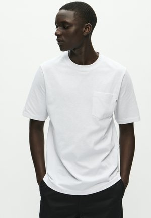 Personne à la peau foncée portant un t-shirt blanc uni à manches courtes avec une poche poitrine, les mains dans les poches, regardant sur le côté contre un fond blanc.