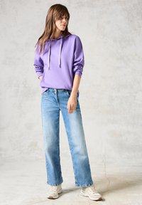 Lila huvtröja med dragsko, kombinerad med ljusblå vida jeans och vita sneakers, stående på en strukturerad bakgrund.
