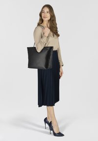 Borsa tote in pelle nera con manici laterali, design minimalista, texture liscia, portata da una persona in blusa beige e gonna plissettata blu navy.