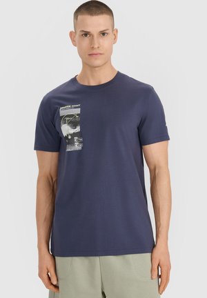 Młody mężczyzna o krótkich włosach, ubrany w granatowy t-shirt z grafiką i jasnobeżowe spodnie, patrzący przed siebie z neutralnym wyrazem twarzy.